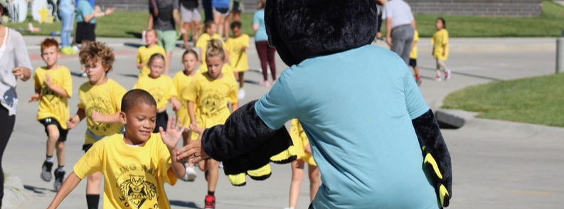 student giving high five to the Guardian mascot while a group of students running in the Fun Run wearing blue Falling Waters Elementary tshirts