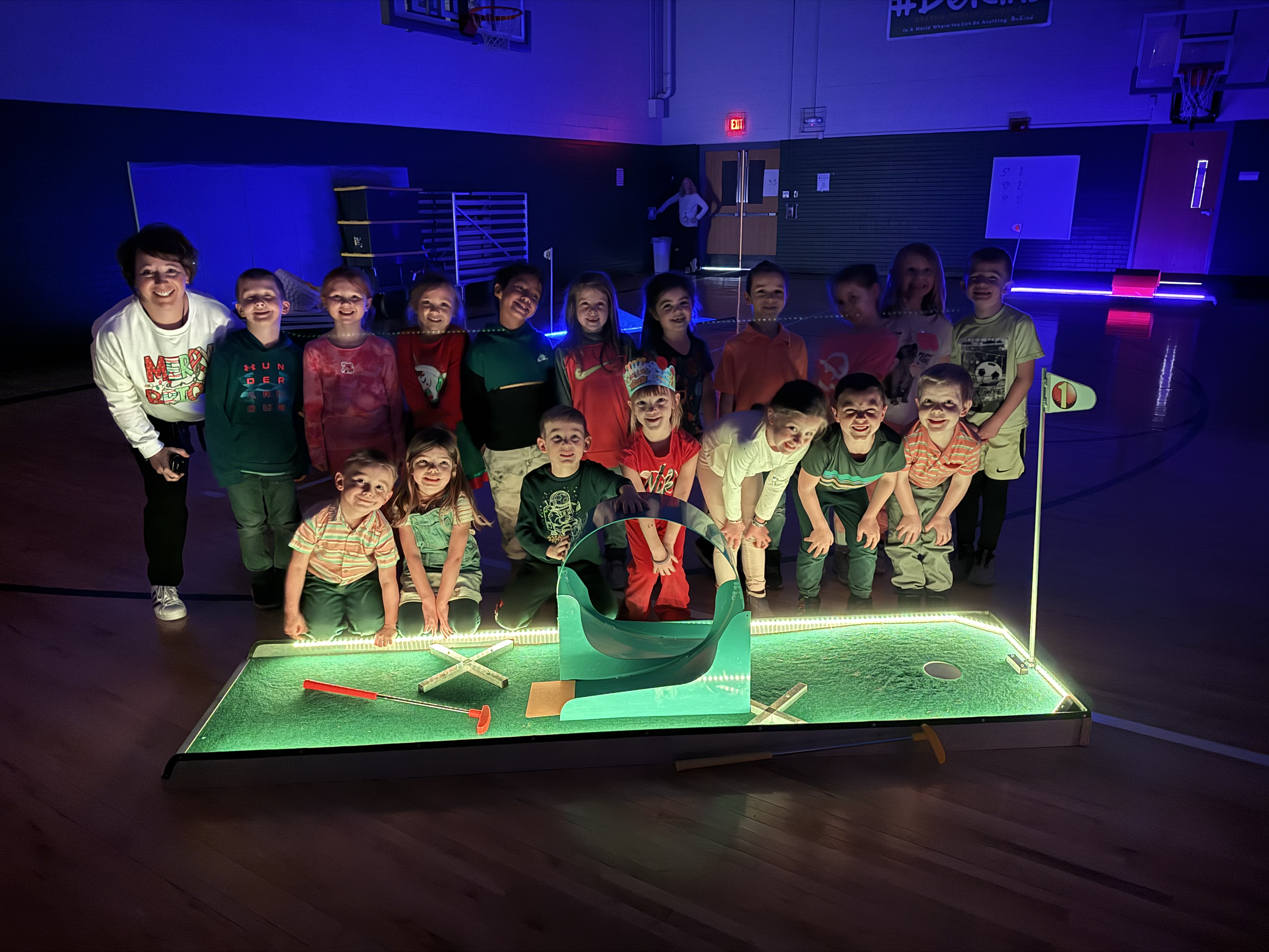 Group of kindergarten students and their teacher posing for a picture in front of a neon lit up mini golf hole with the lights turned off in a gymnasium.