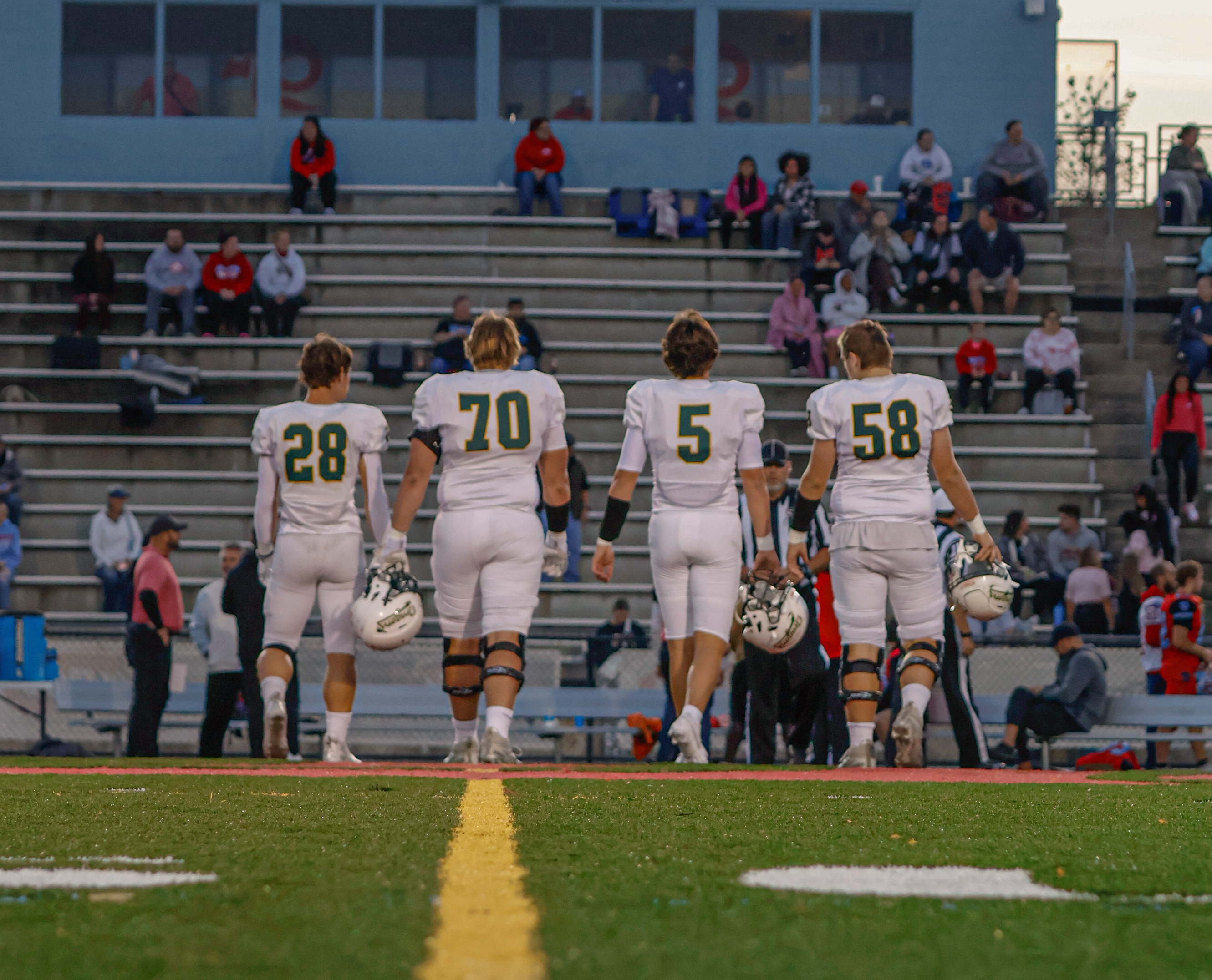 four football players walking away from the camera to the sideline with helmets off and holding at side.