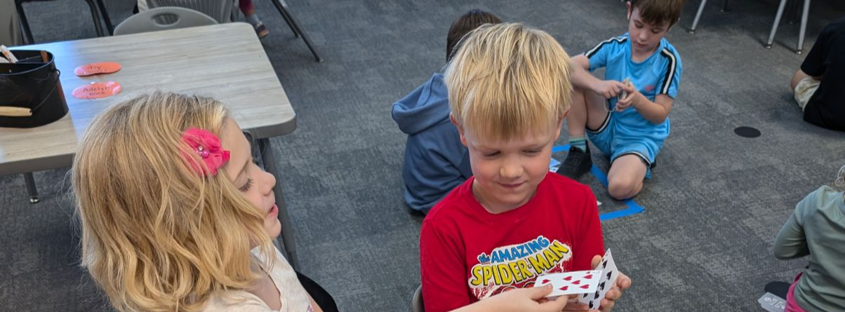 two young students playing cards together in the classroom, other students in the background doing the same