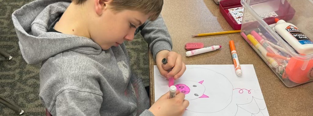 Student drawing and coloring a pig at classroom desk.