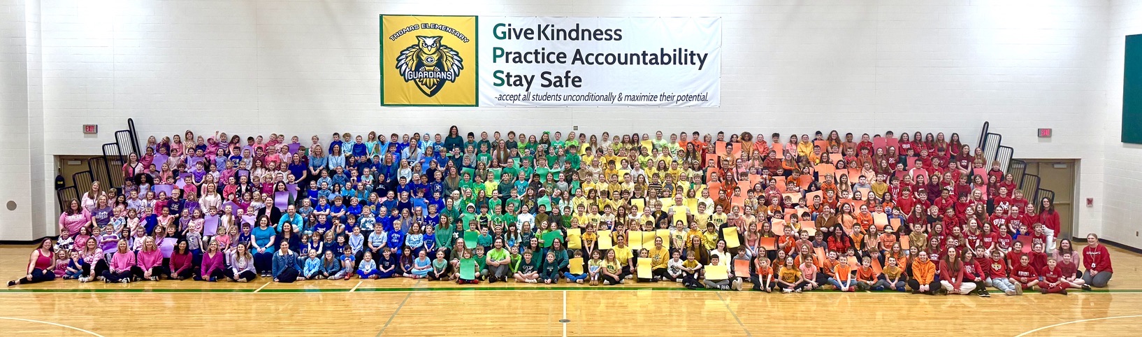 Thomas Elementary Students seated in the gym with their colors of the rainbow shirts on for each grade level. 