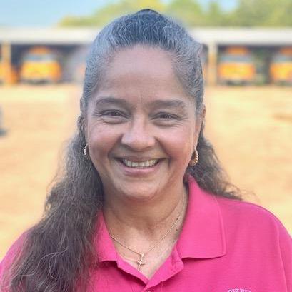 A smiling woman in a pink shirt standing in front of a school bus.
