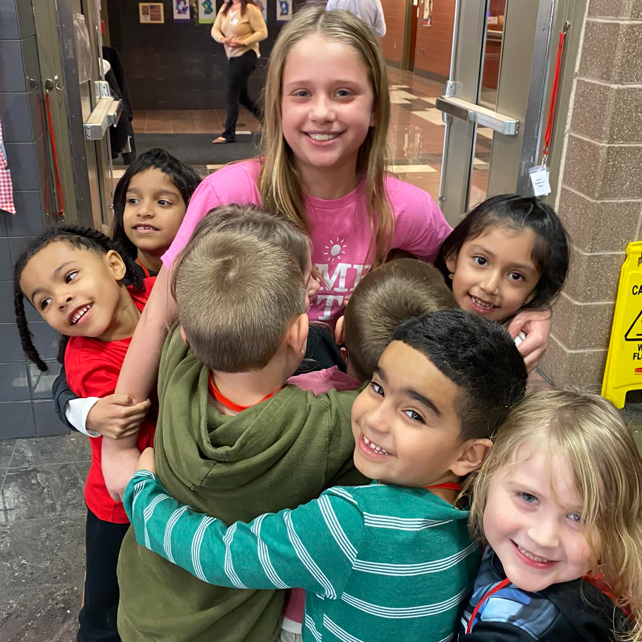 Group of smiling children hugging older student in colorful shirt.