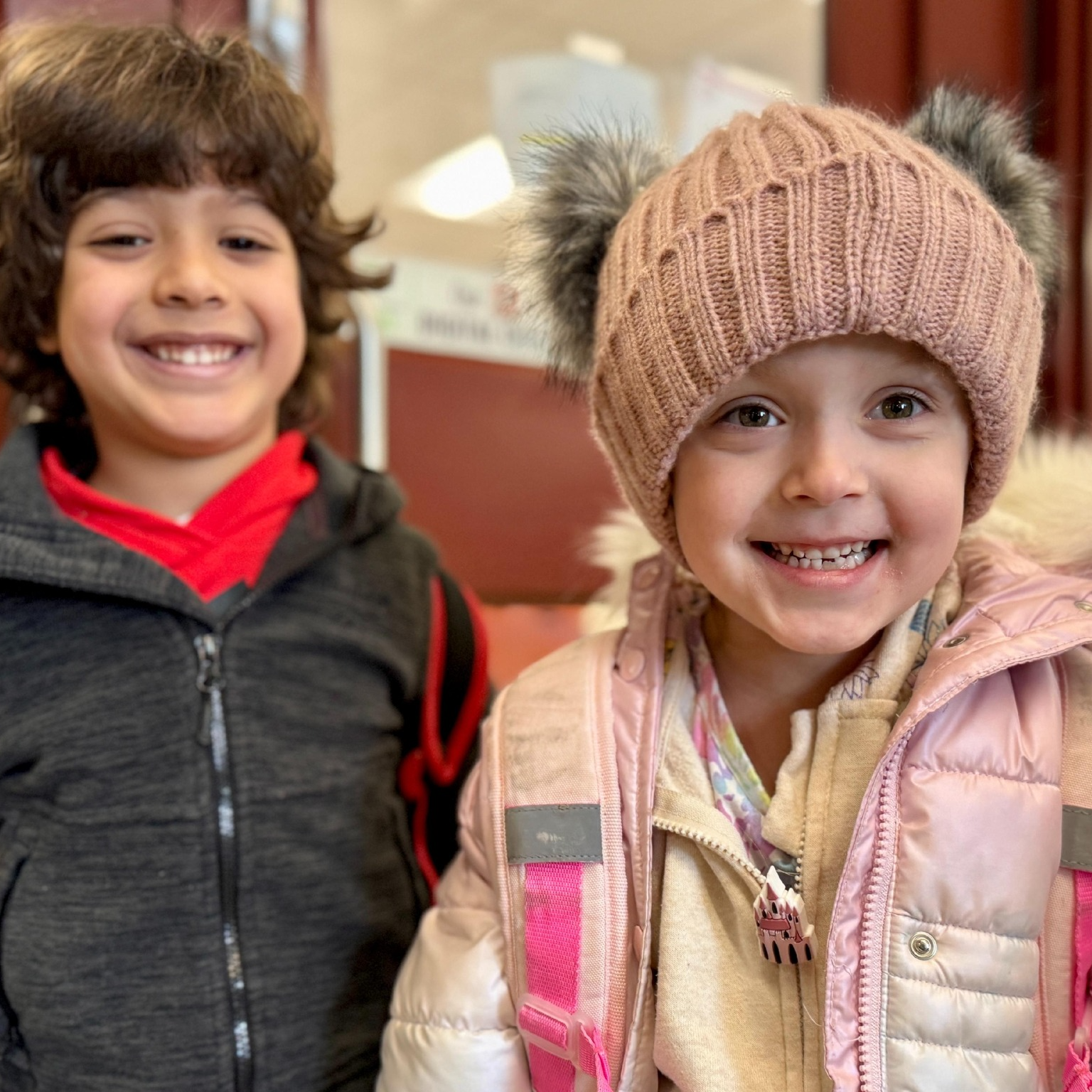 Young girl in jacket and pink hat and young boy in gray sweatshirt. Both are smiling.