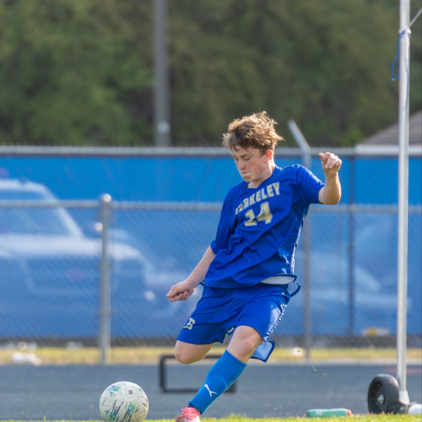 A boy in a blue uniform kicks a soccer ball on a grassy field with a fence in the background.