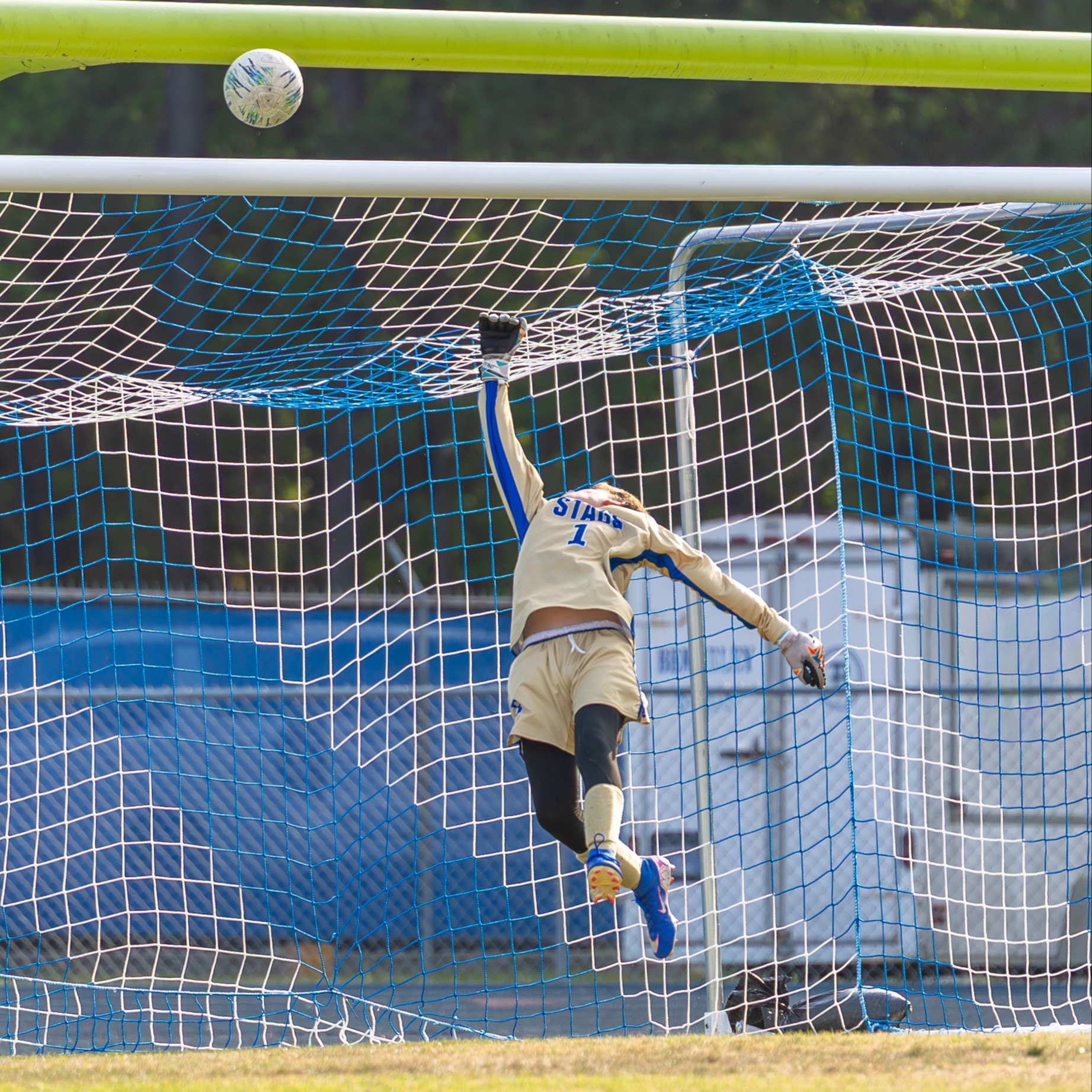A soccer player leaps to block a ball in mid-air, positioned in front of a soccer goal.