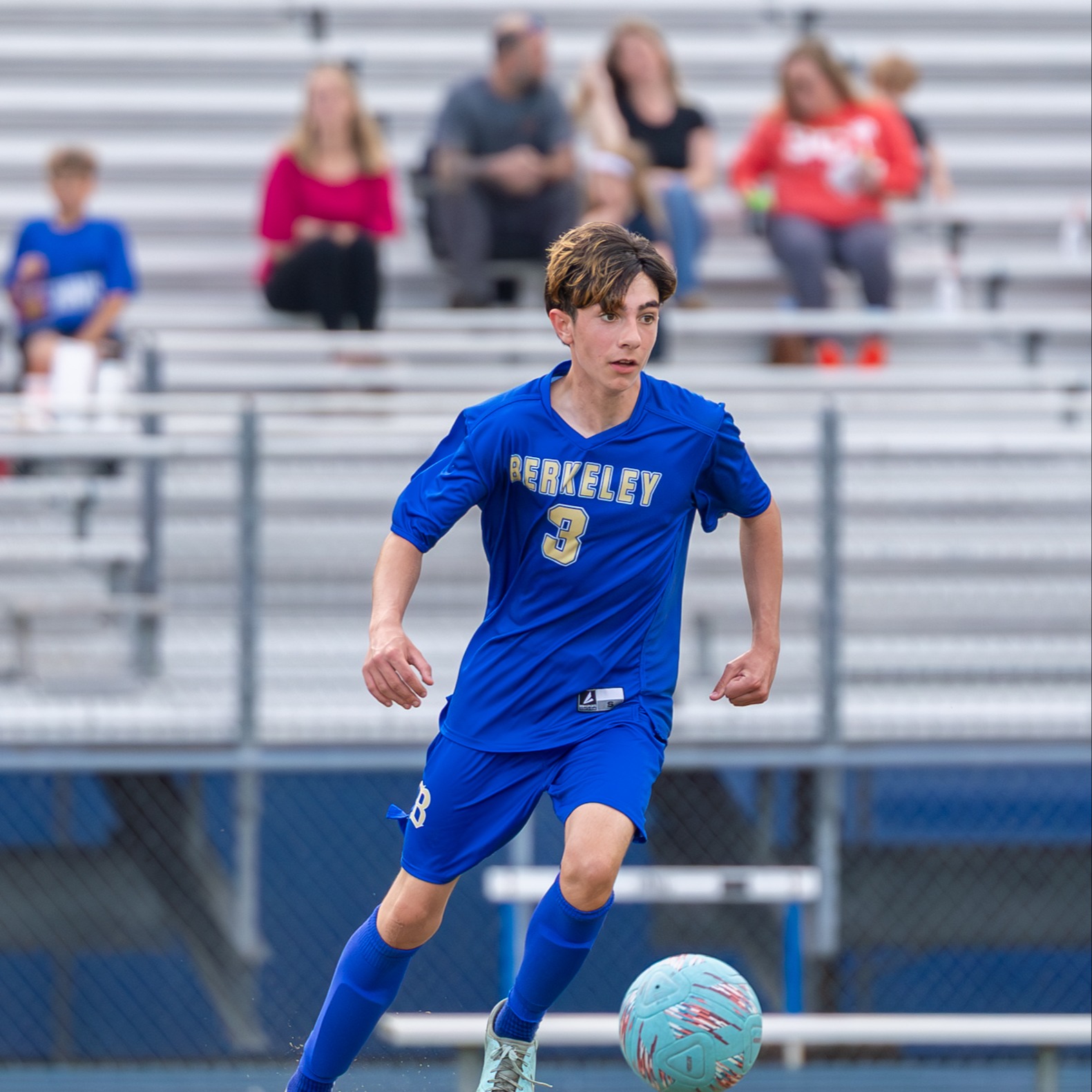 A boy in a blue jersey runs towards a soccer ball on a field with spectators seated on bleachers.