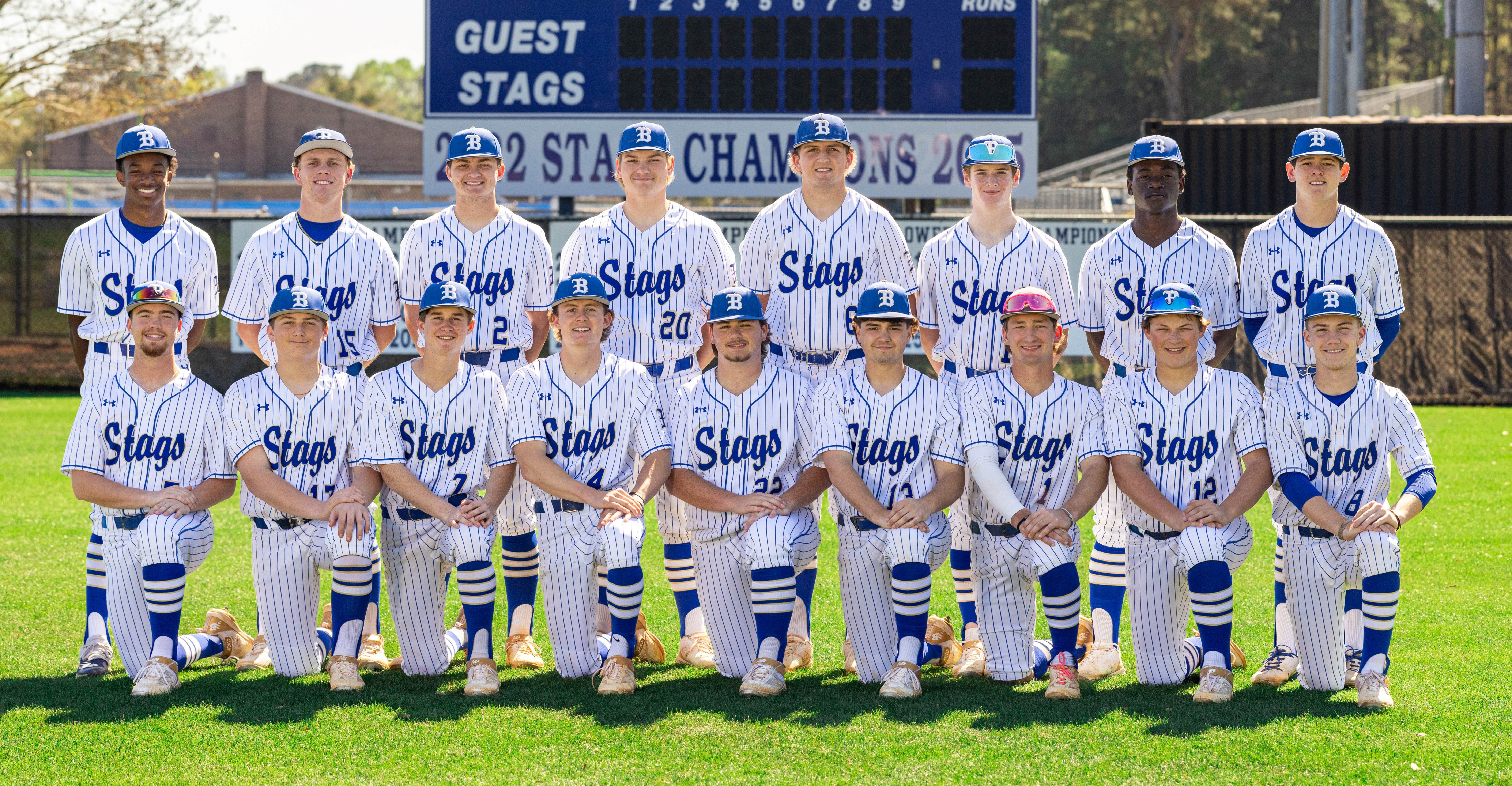 A baseball team, in white and blue uniforms, poses for a team photo in front of a scoreboard.