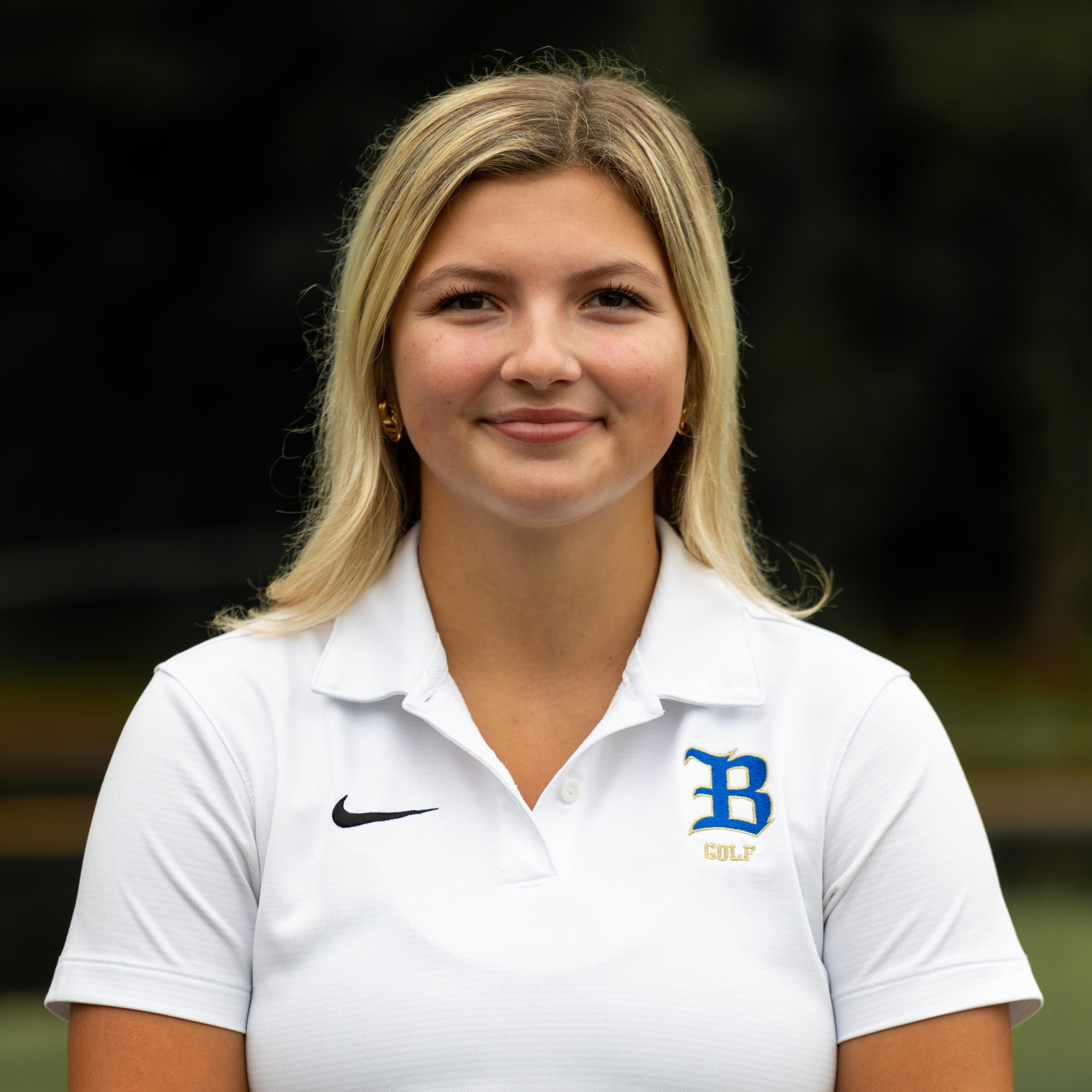 Woman with blonde hair wearing white shirt with Nike and BU logo, smiling, standing in a park.