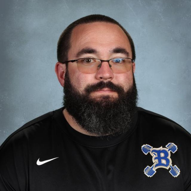 A man with a beard wearing glasses and a black shirt with a logo on the chest.