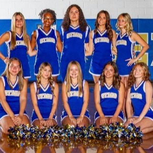 A group of female cheerleaders in blue uniforms with white letters, some kneeling with pom-poms, posing in a gym.