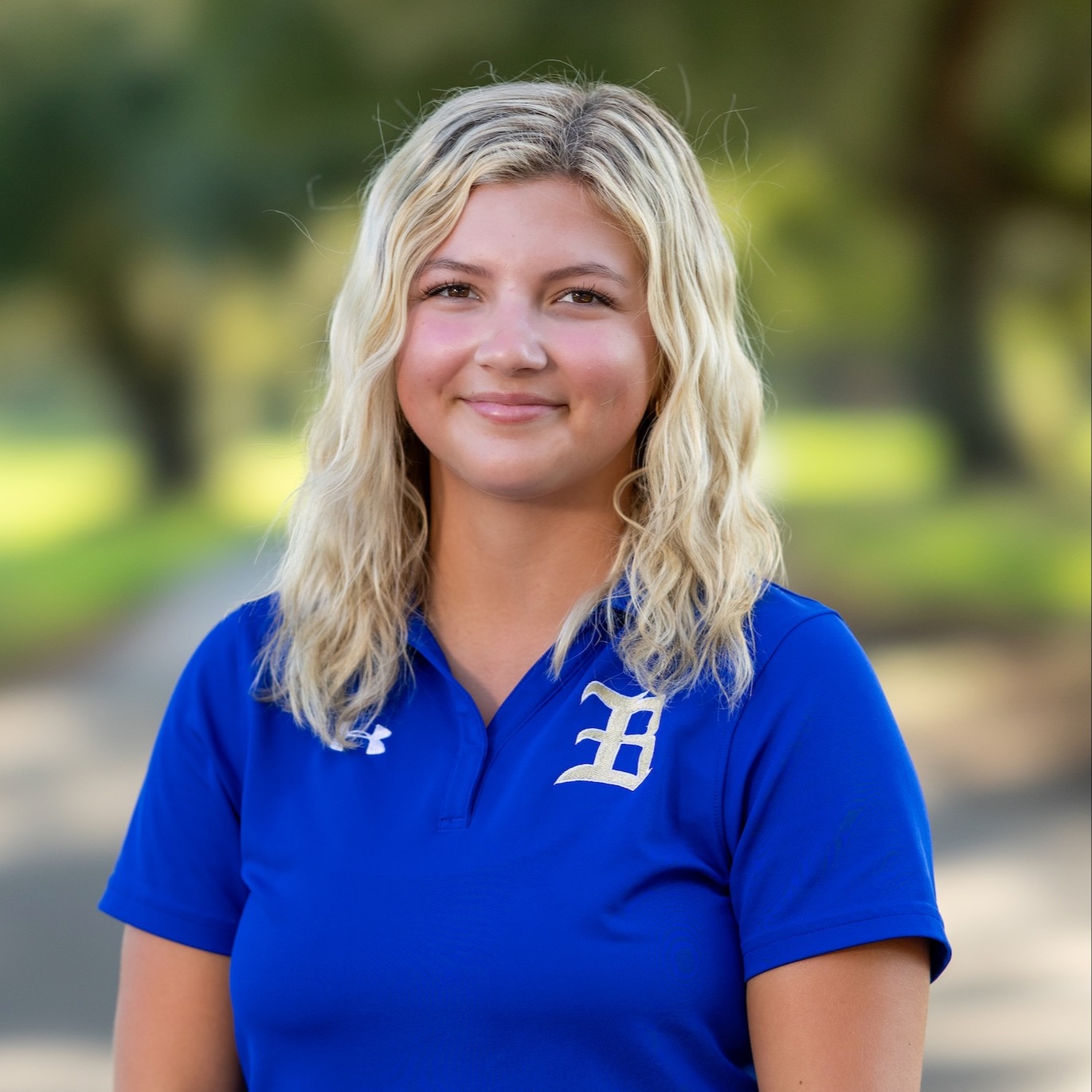 A woman with blonde hair stands outside, wearing a blue polo with the letter "D" on it.