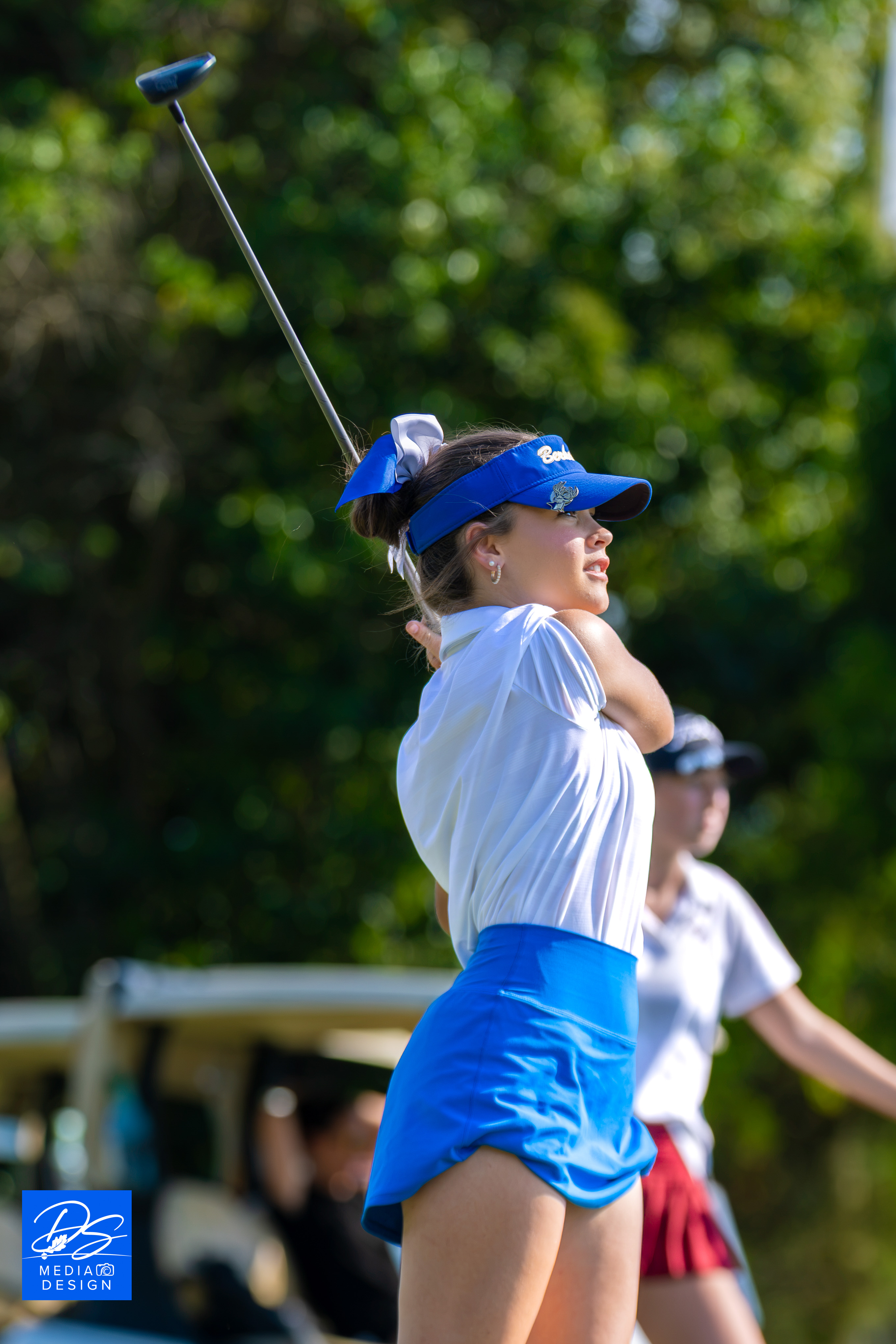 A woman in a white shirt and blue visor swings a golf club on a golf course.