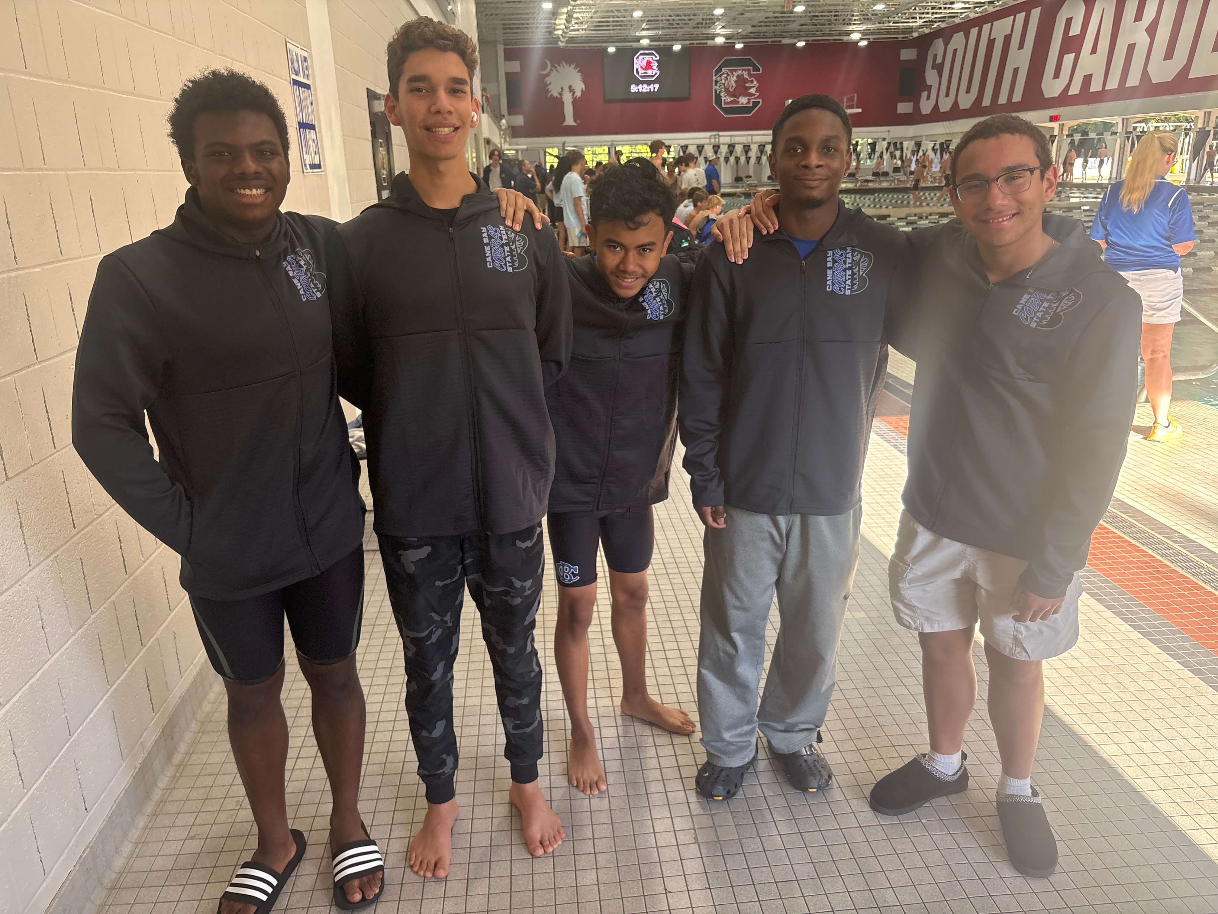 Five young males standing together in a tiled corridor, wearing black and gray athletic wear. Behind them, a wall reads "South Car."