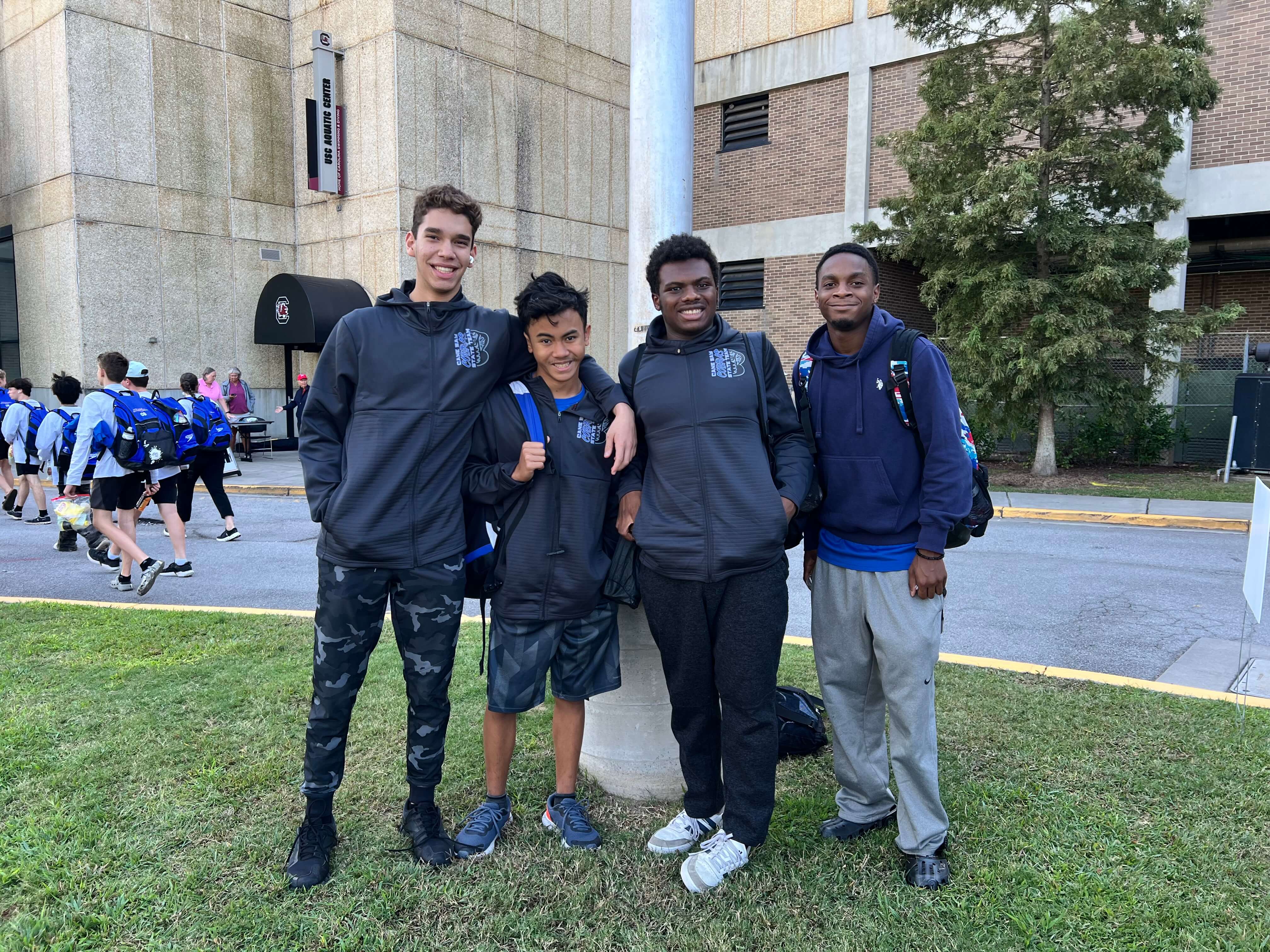 Four individuals stand together on grass with a building, trees, and people walking in the background.