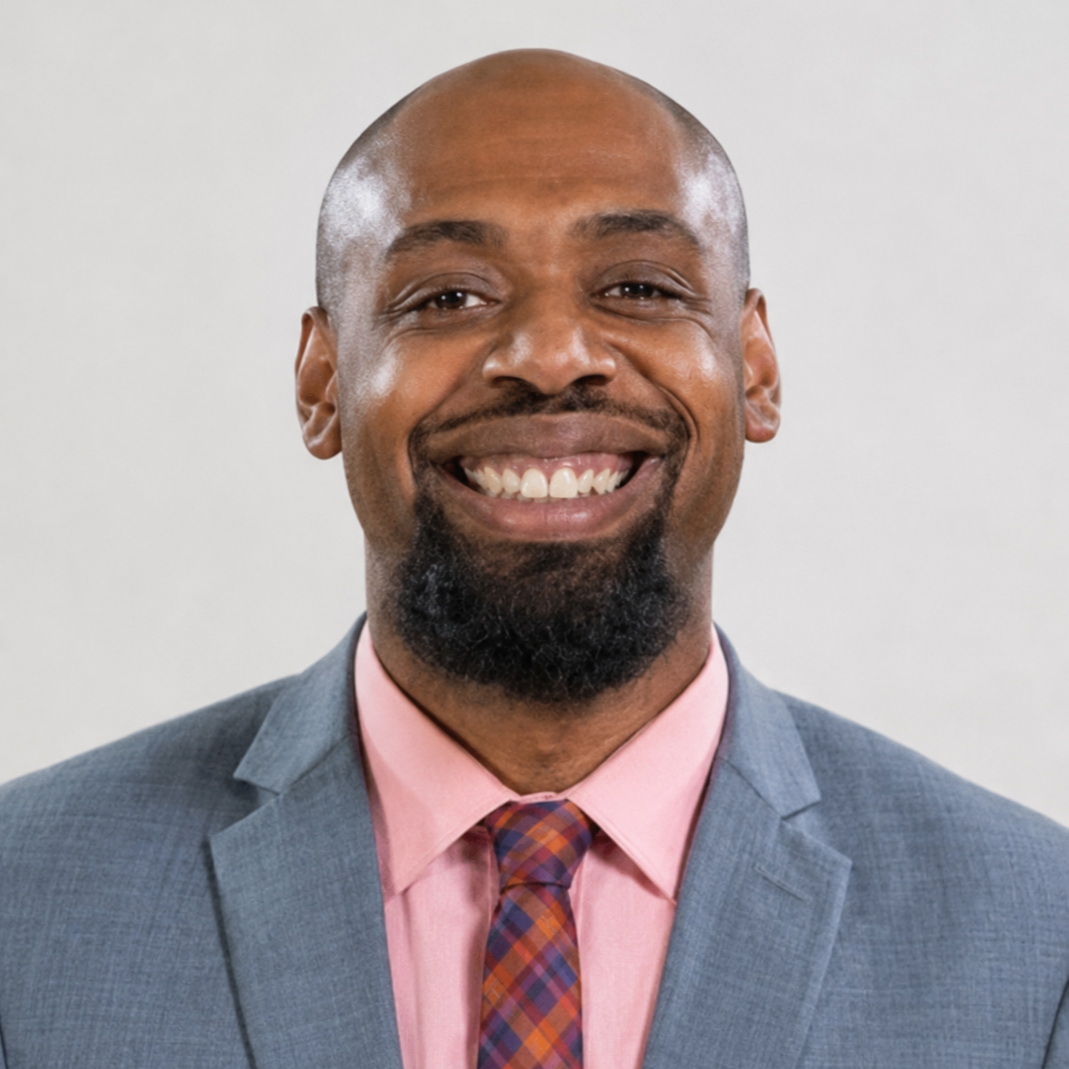 Man smiling, wearing a gray suit jacket, pink shirt, and tie with orange, blue, and white squares.