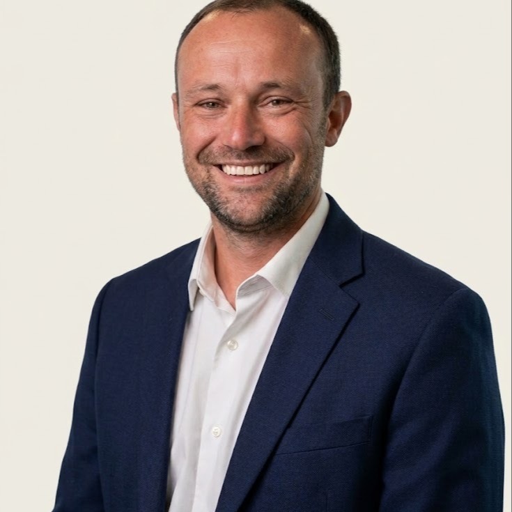 A smiling man in a white shirt and navy blazer poses for a photo against a plain background.