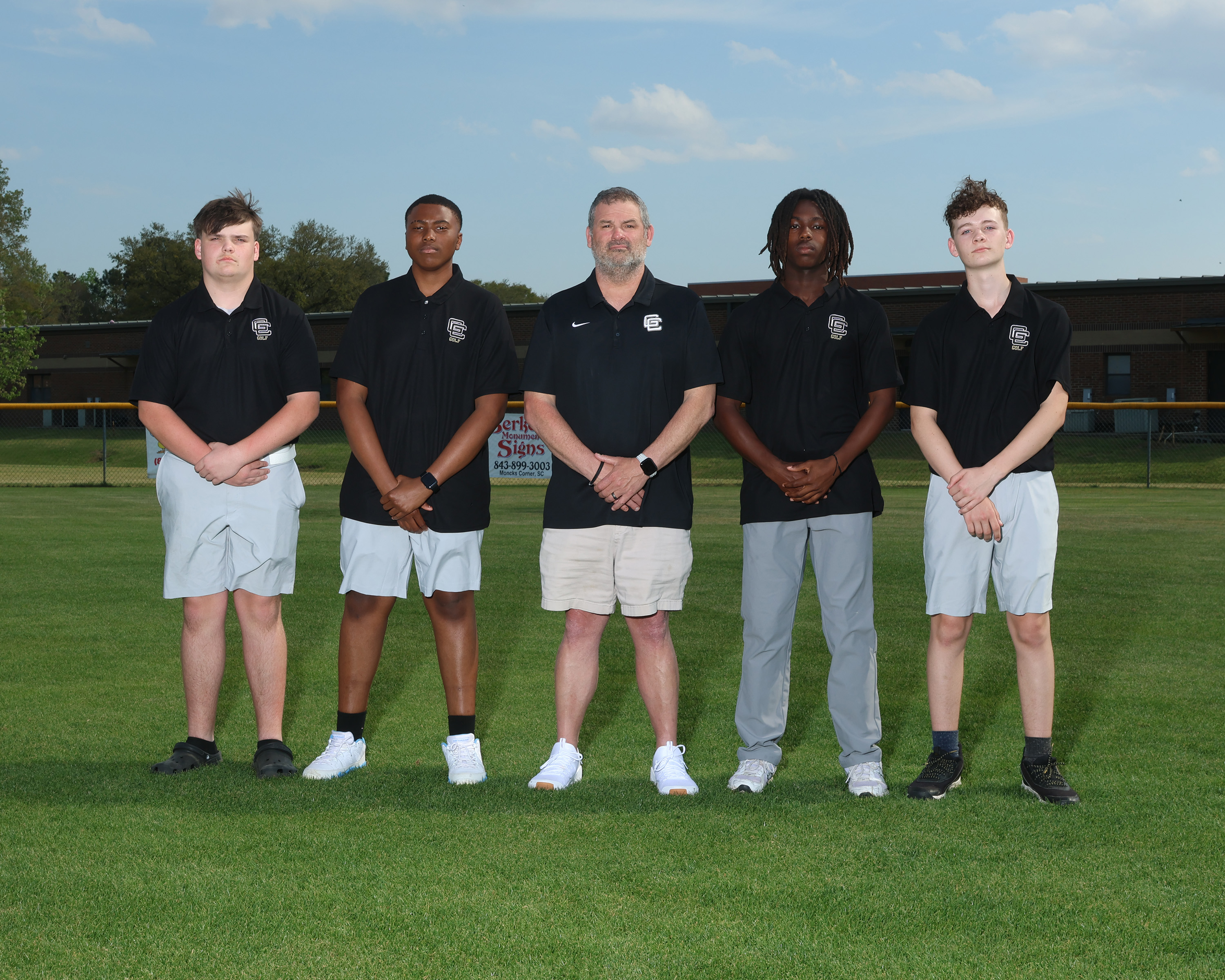 Six individuals in matching black shirts and shorts pose on a green field with a baseball diamond in the background.