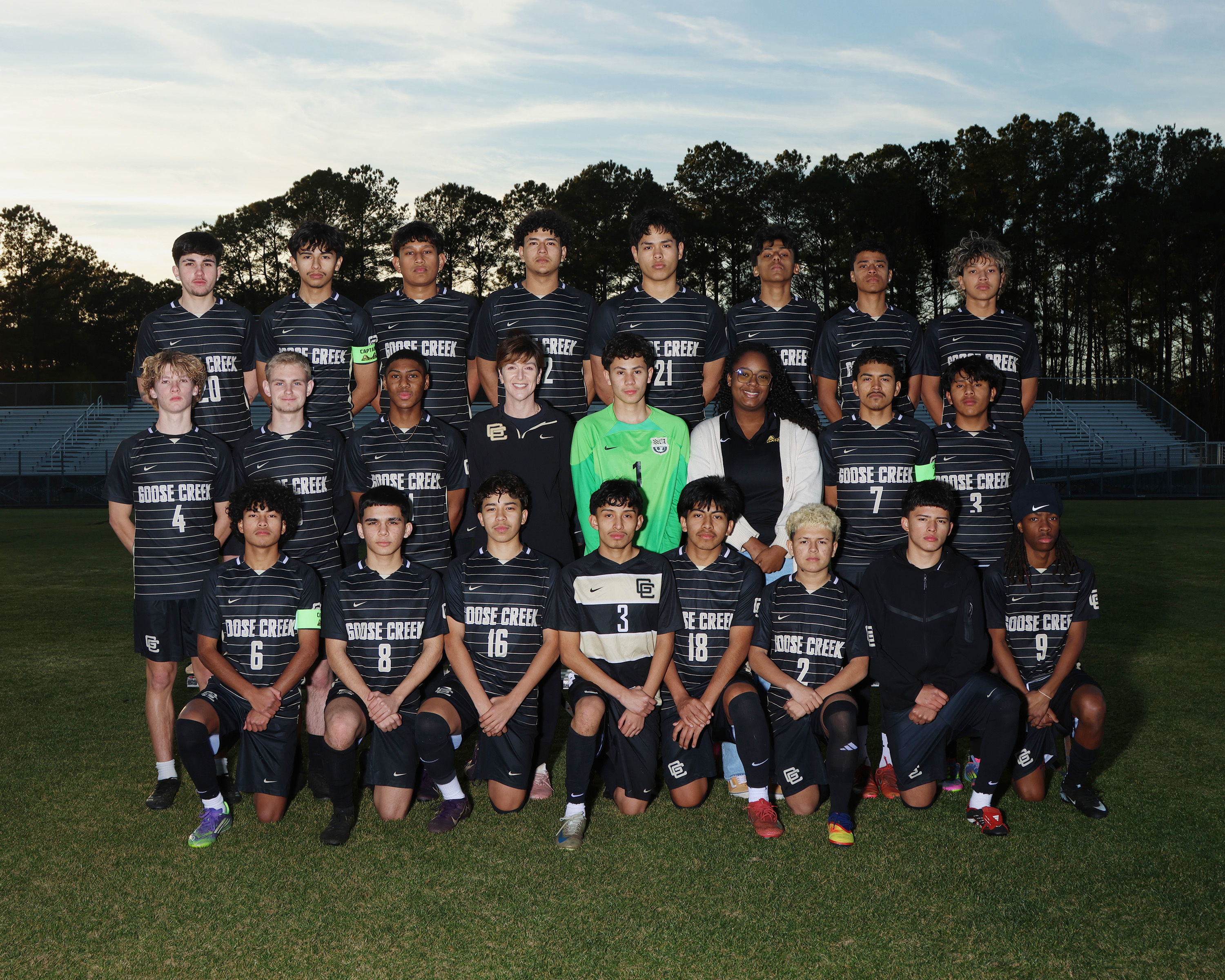 A soccer team stands on a field in two rows. They wear black uniforms with white stripes. Behind them, trees line the horizon.