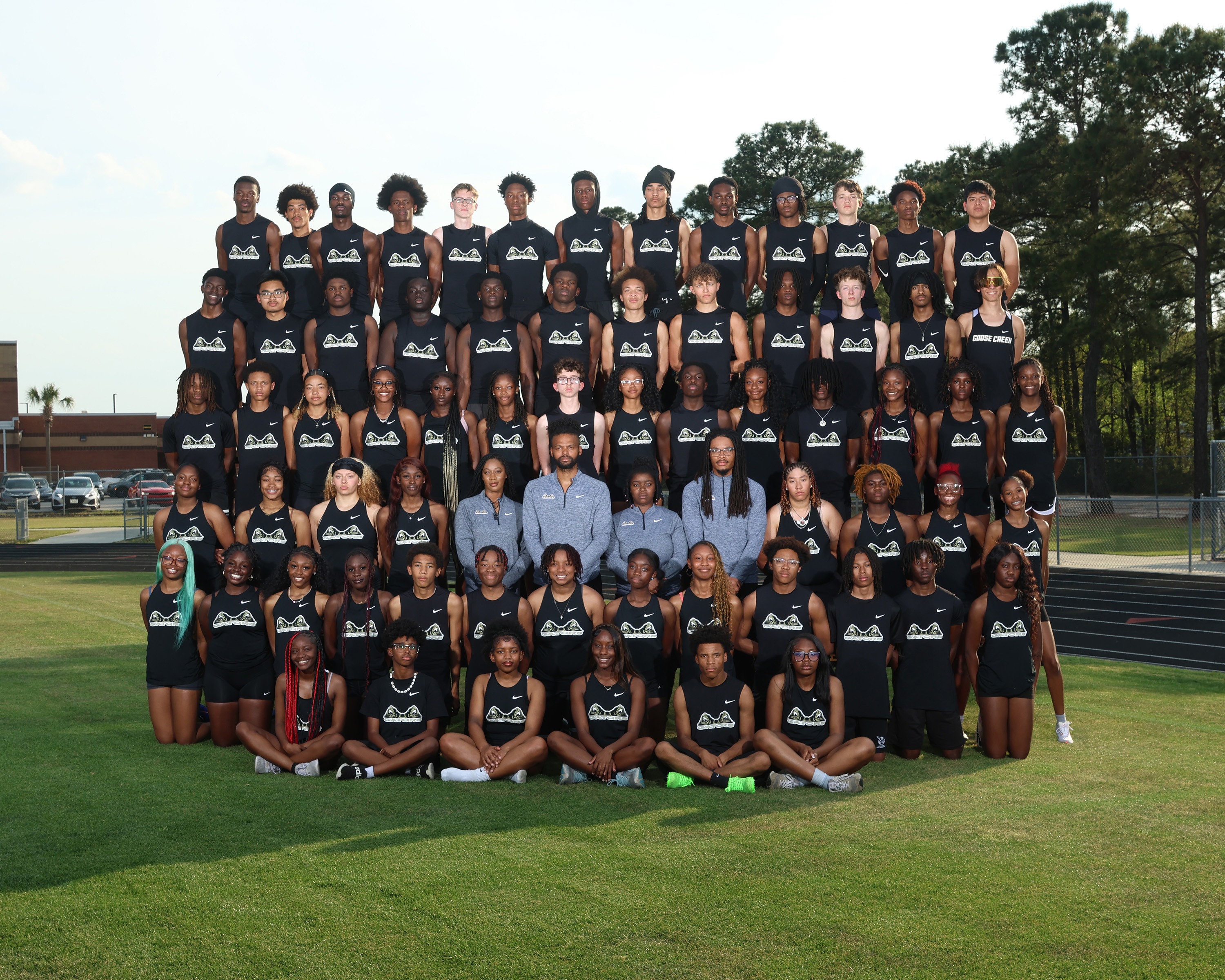 Group photo of a sports team, all in black uniforms, posing on a grassy field with trees and a track in the background.