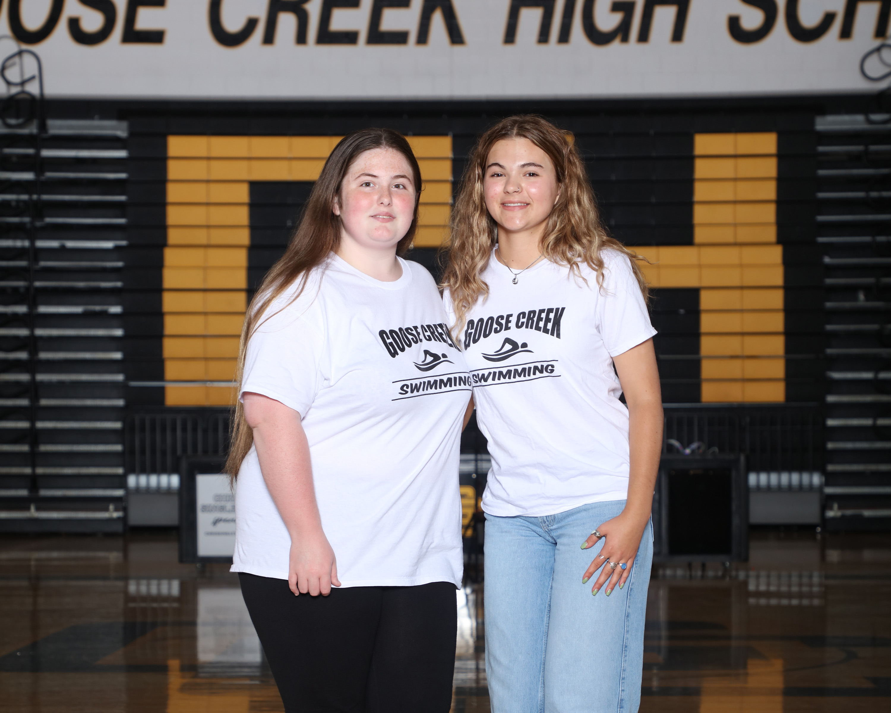 Two women, wearing white shirts, stand side by side in a gymnasium. Behind them is a wall with a "Rose Creek High School" banner.