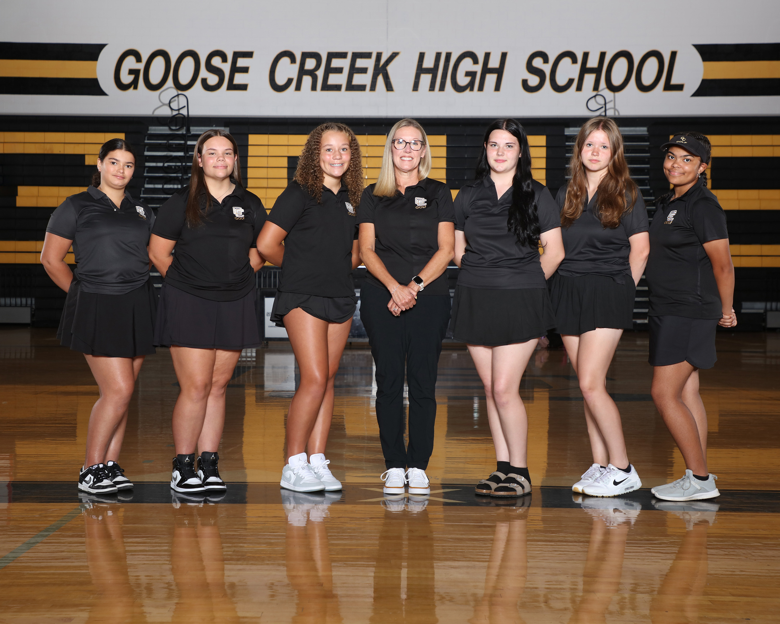 Nine individuals in matching uniforms stand on a basketball court. A sign behind them reads "GOOSE CREEK HIGH SCHOOL."