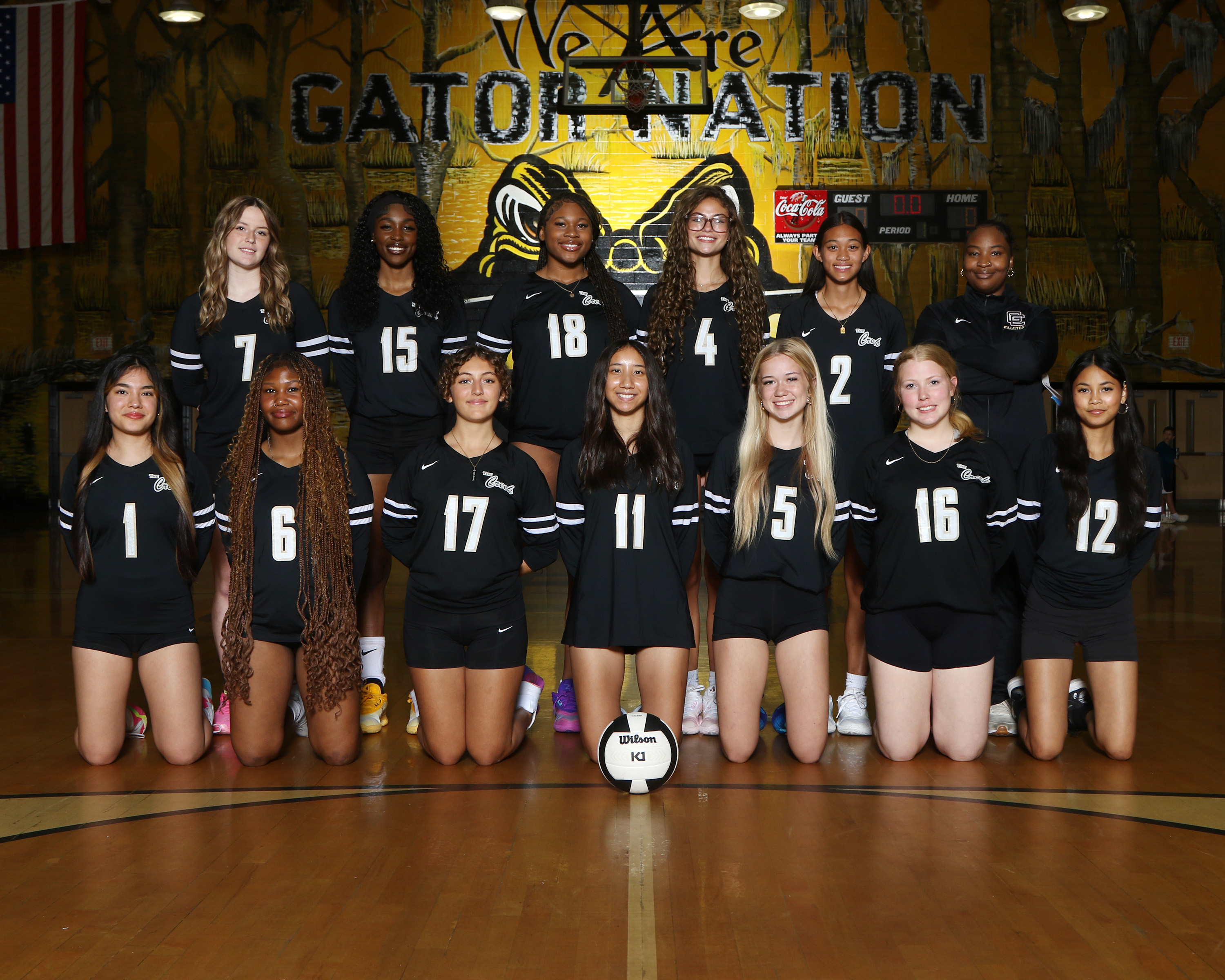 A volleyball team in black uniforms poses on a court with a ball. Behind them, a wall displays "We Are Gator Nation".