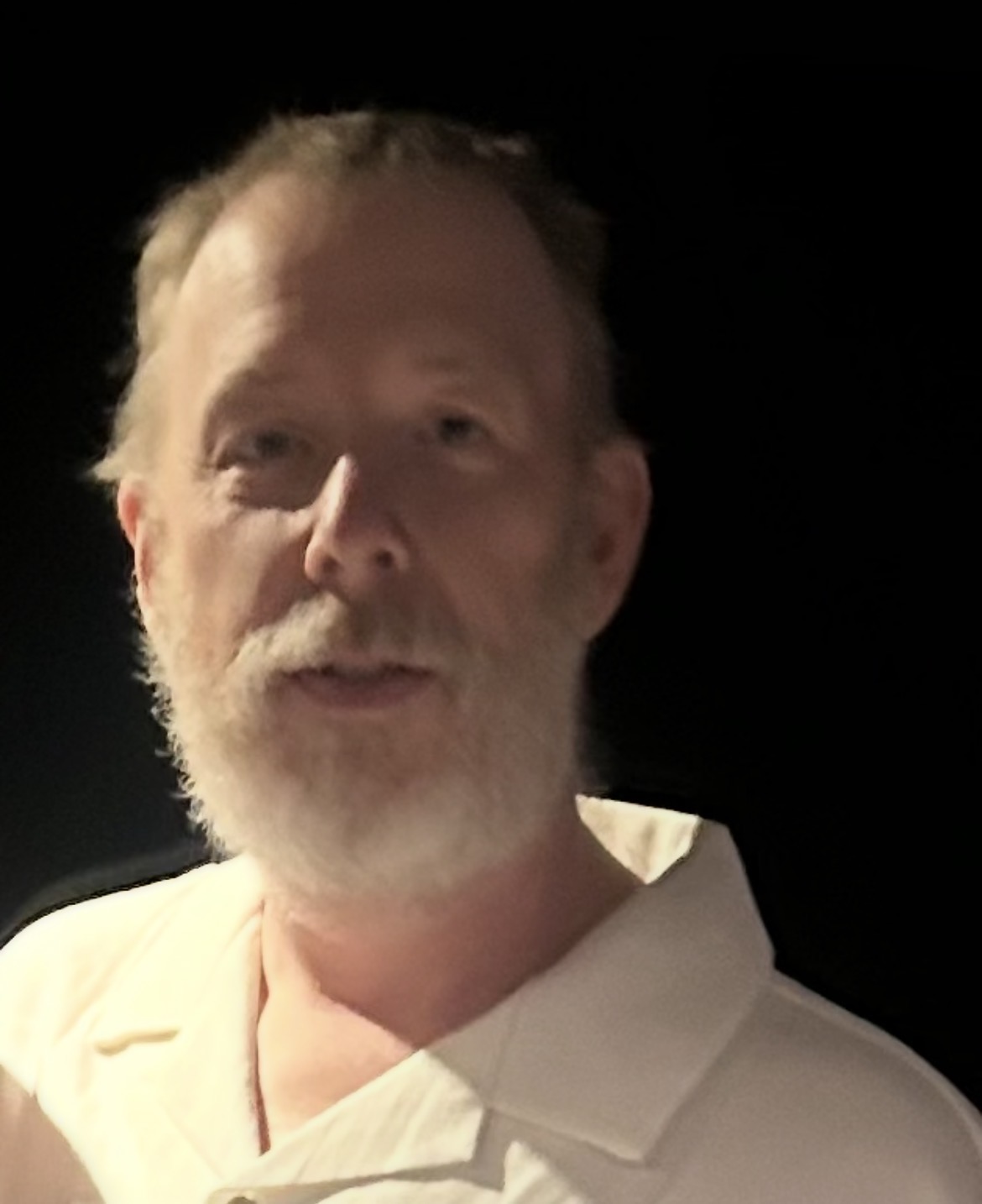 A man with gray hair and a white beard wearing a white polo shirt stands in front of a dark background.