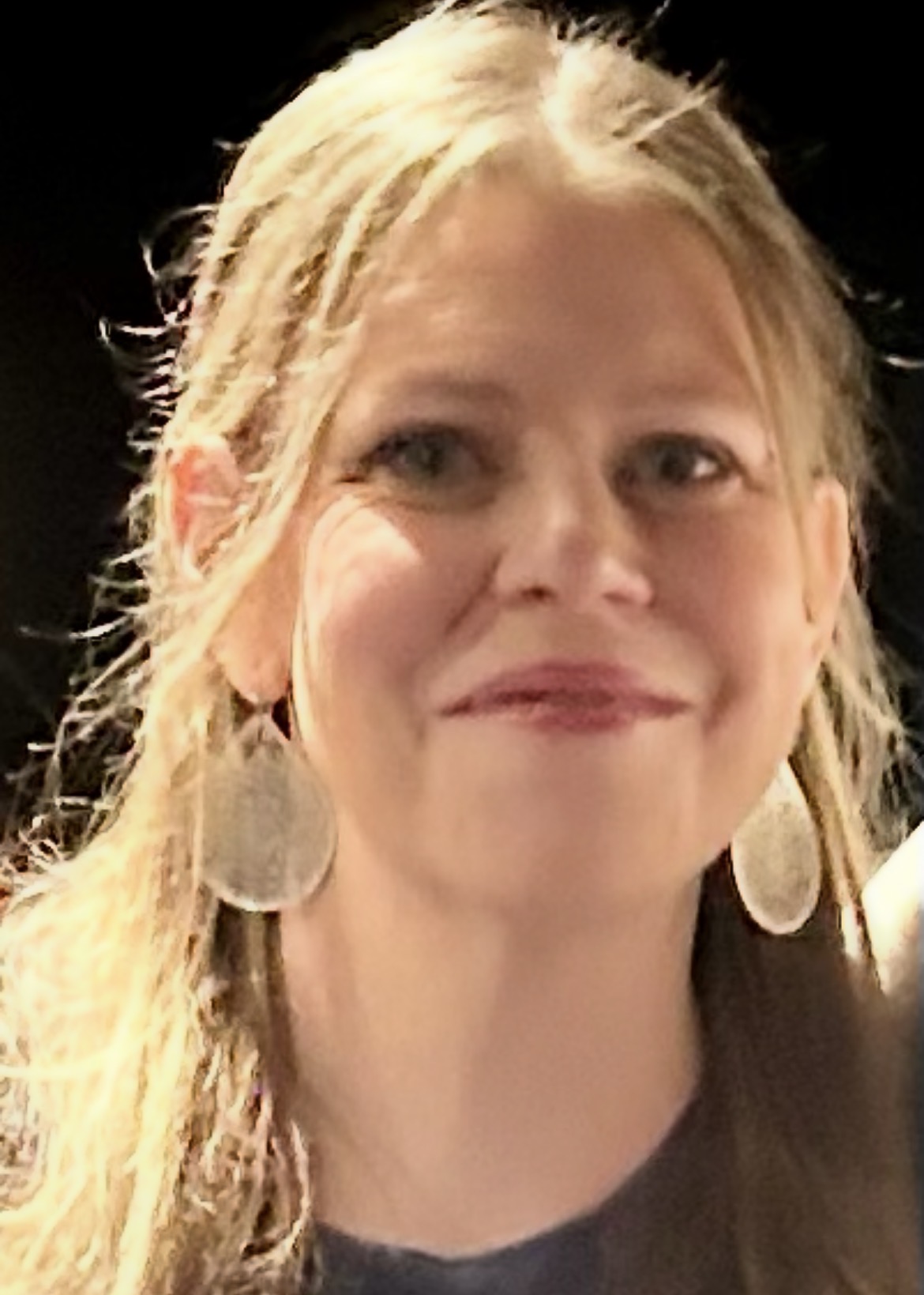 A woman with long blonde hair wearing a gray shirt and large silver earrings smiles at the camera.