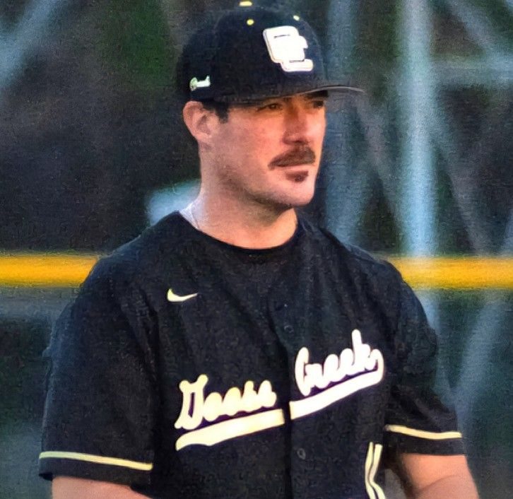 A baseball player stands on a field, wearing a black jersey with yellow and white text.