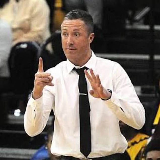 Coach giving instructions on a basketball court with two men and spectators in the background.