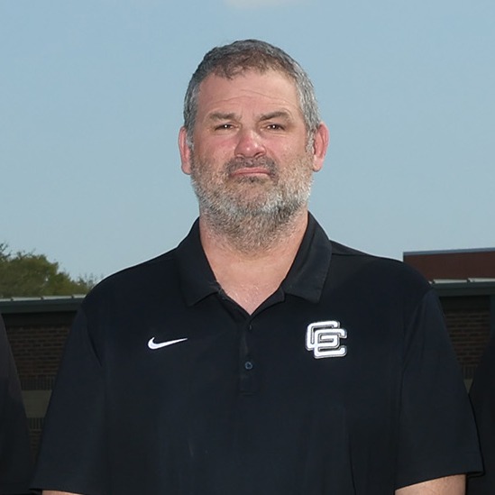 Six individuals, wearing black shirts, stand on a green field. They are positioned closely together.