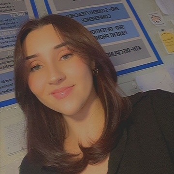 A woman with brown hair and a black top stands in front of a white wall with posters.