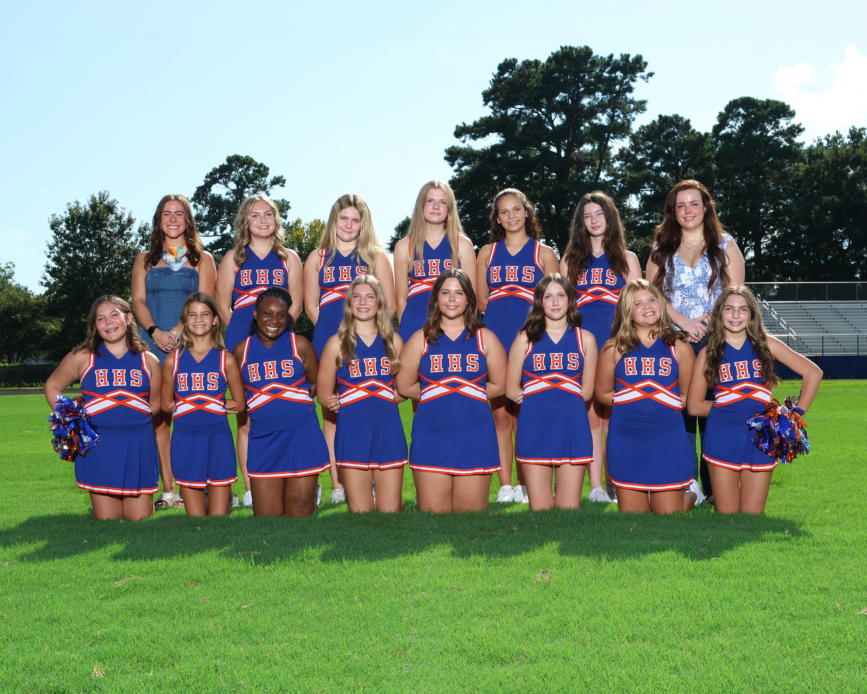 A group of students dressed in blue cheerleading uniforms posing on a grass field with trees.