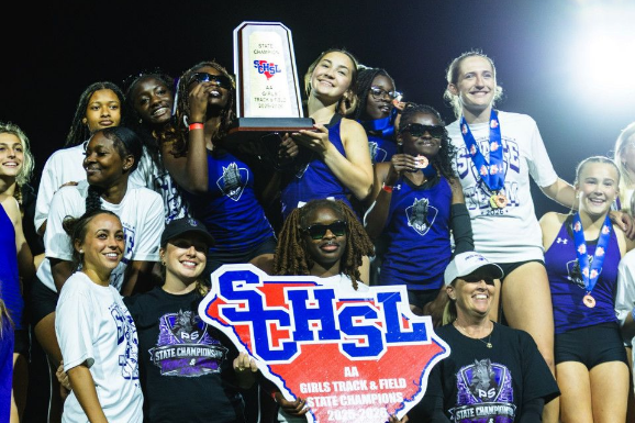 A group of women holding a large plaque, smiling, and posing for a photo on a sports field.