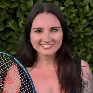 Woman with tattoos holding tennis racket, smiling. Behind her is a green bush.