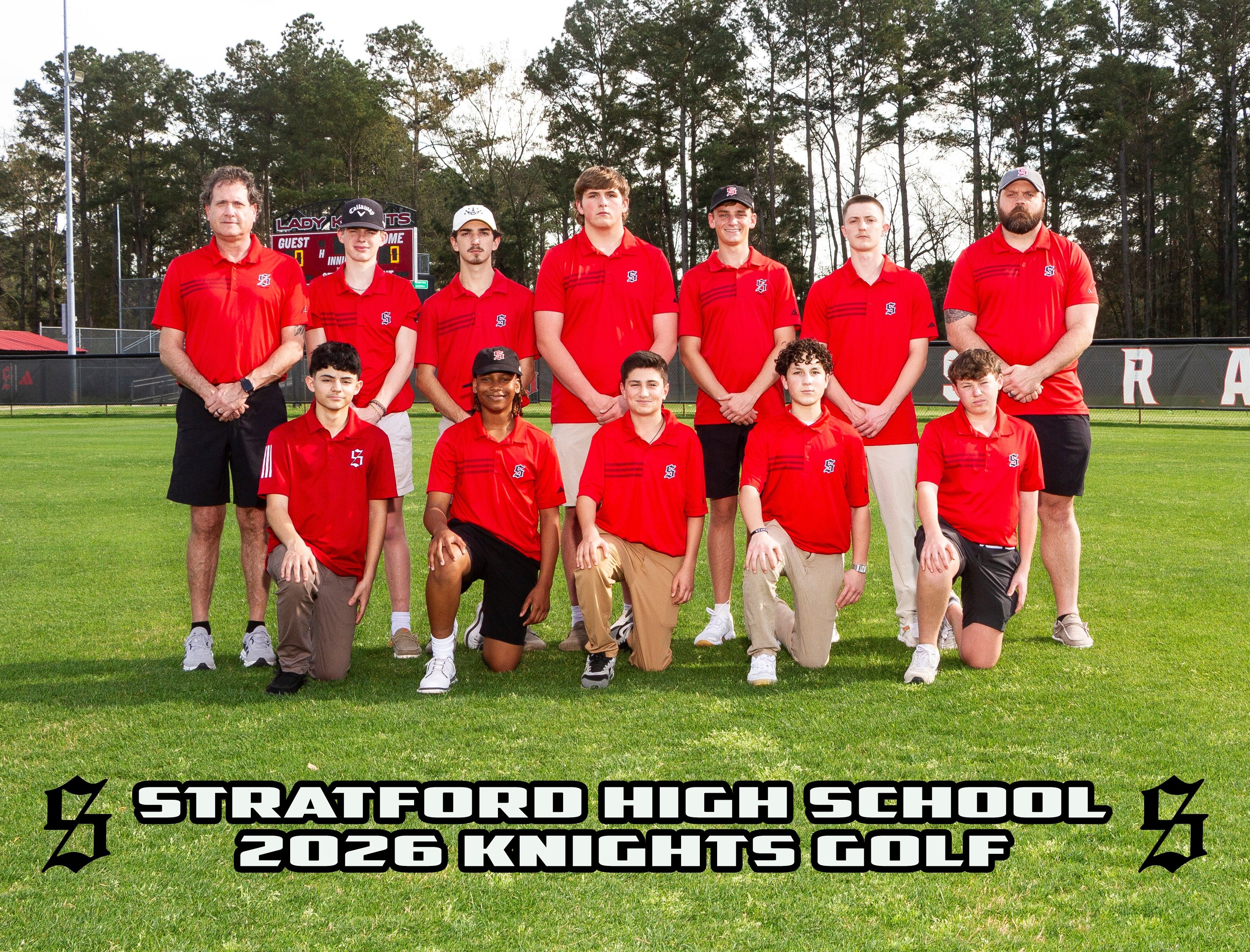 A group of people in red shirts and black shorts pose on a golf course, with trees in the background.