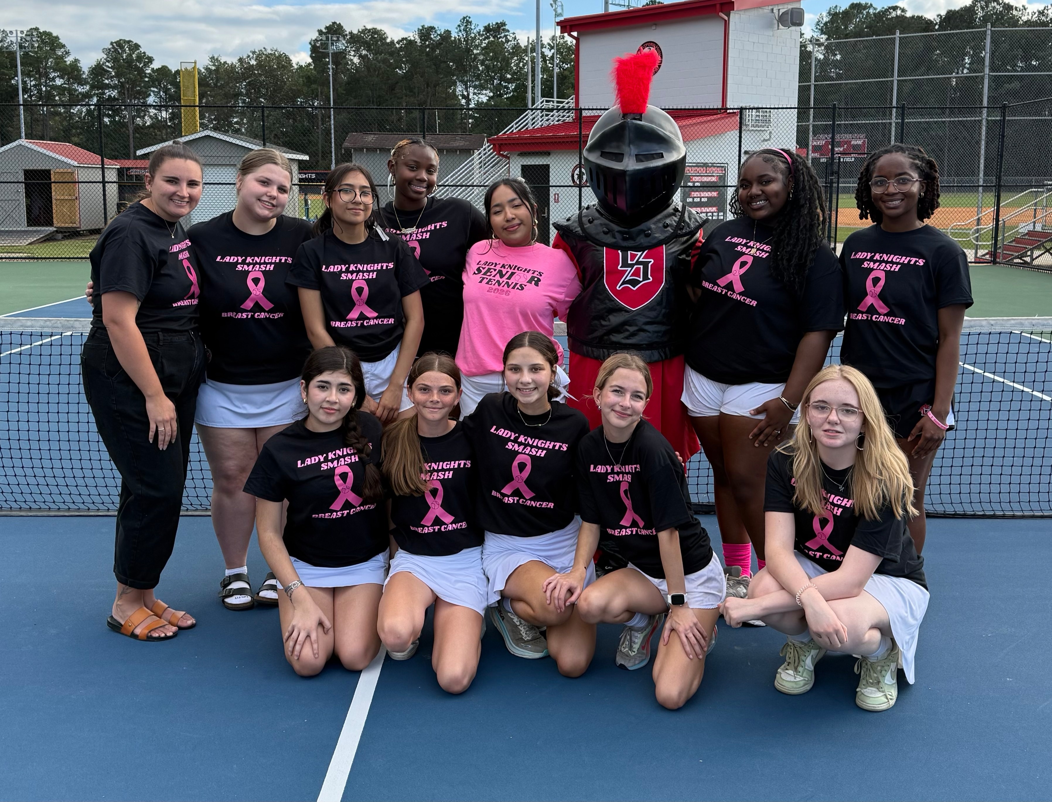 A tennis court with a mascot and a group of people wearing black and pink shirts and shorts.