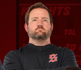 A man stands with arms crossed in front of a red backdrop, wearing a black shirt with a red symbol.