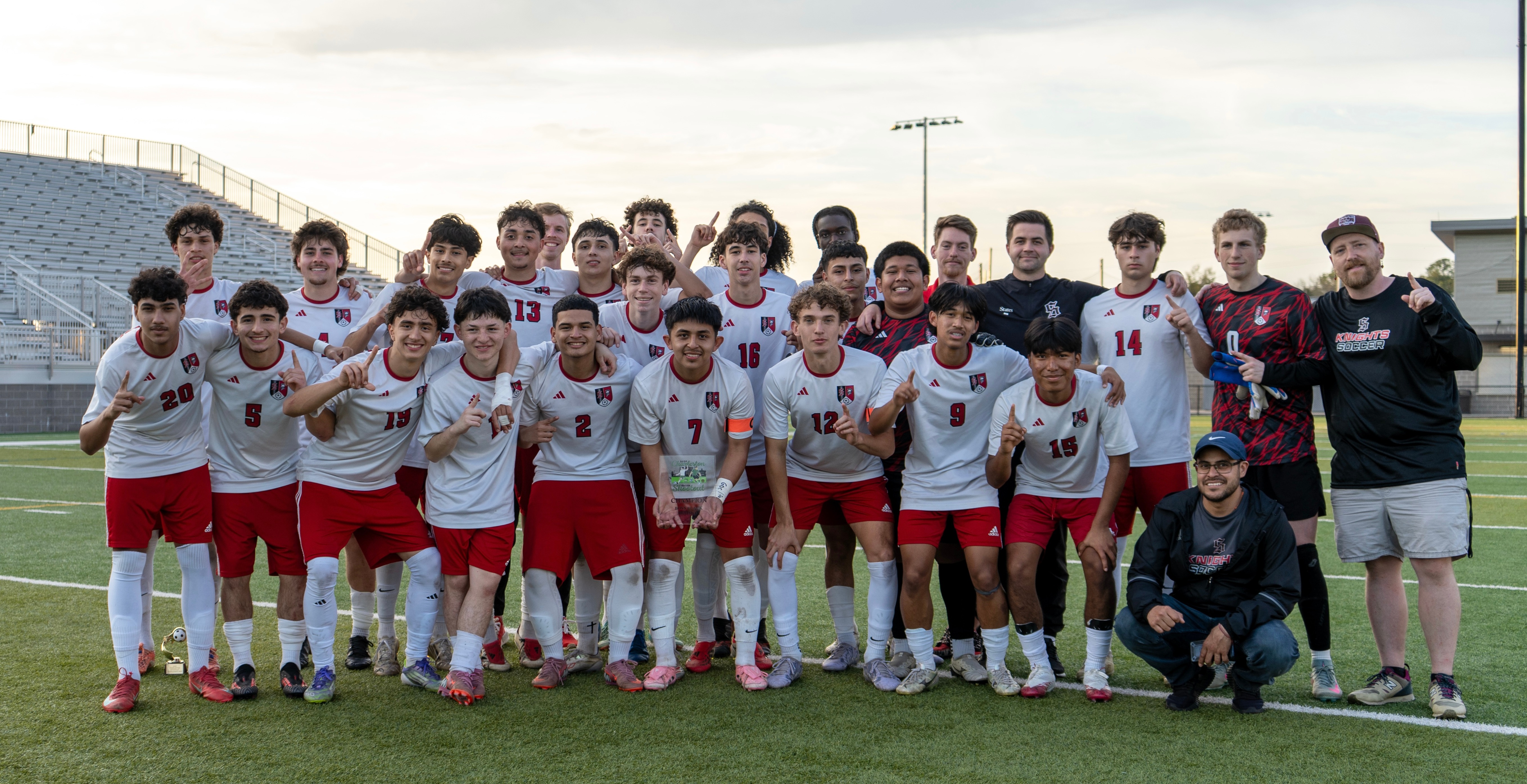 A sports team poses for a photo on a field. The background shows an empty stadium with bleachers and a cloudy sky.
