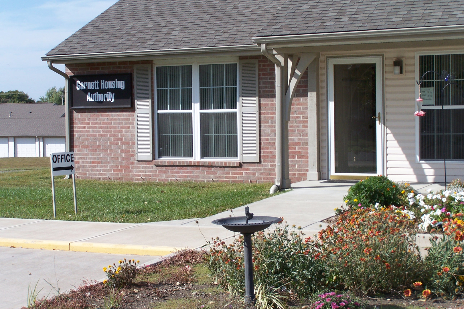 A brick house with a bird bath in front. A sign mounted on the house reads 'Garnett Housing Authority and "OFFICE" below.