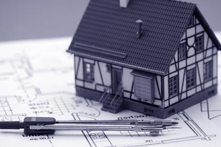 A miniature model of a house with a pitched roof sits on top of a large, unfolded blueprint. The blueprint shows detailed illustrations of the house, including walls, doors, and windows.