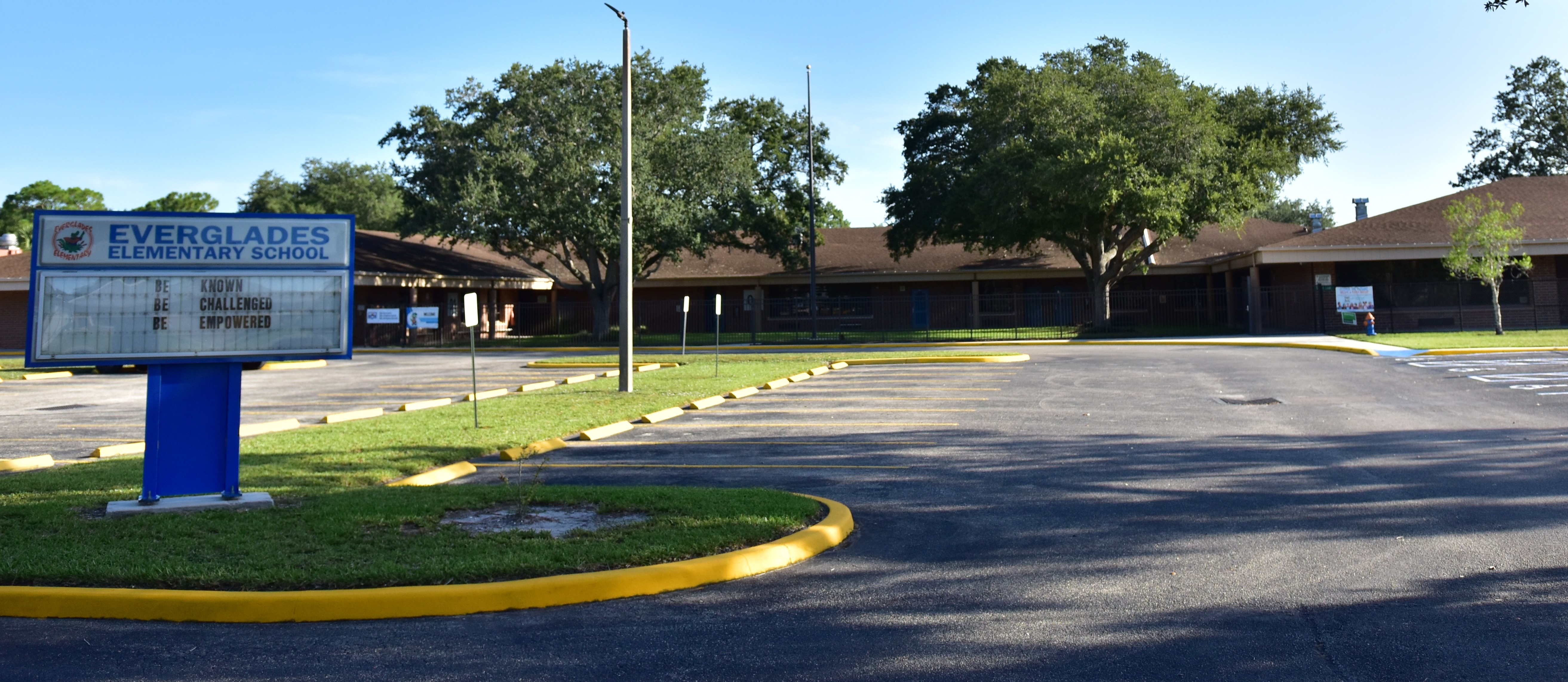 An empty parking lot with a bulletin board and trees in the background. A building is visible behind the trees.