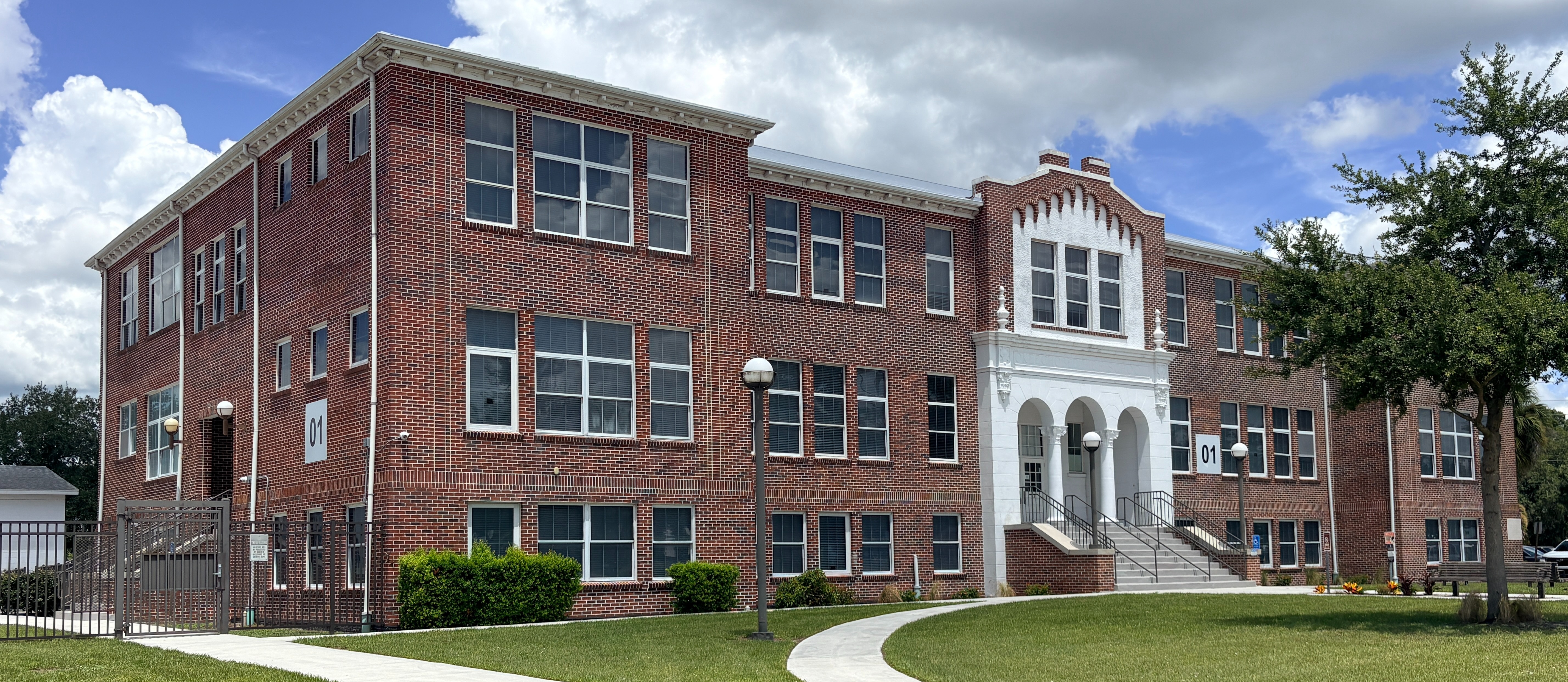 Large brick building with multiple windows and a white entrance. A grassy lawn and sidewalk lead to the entrance.