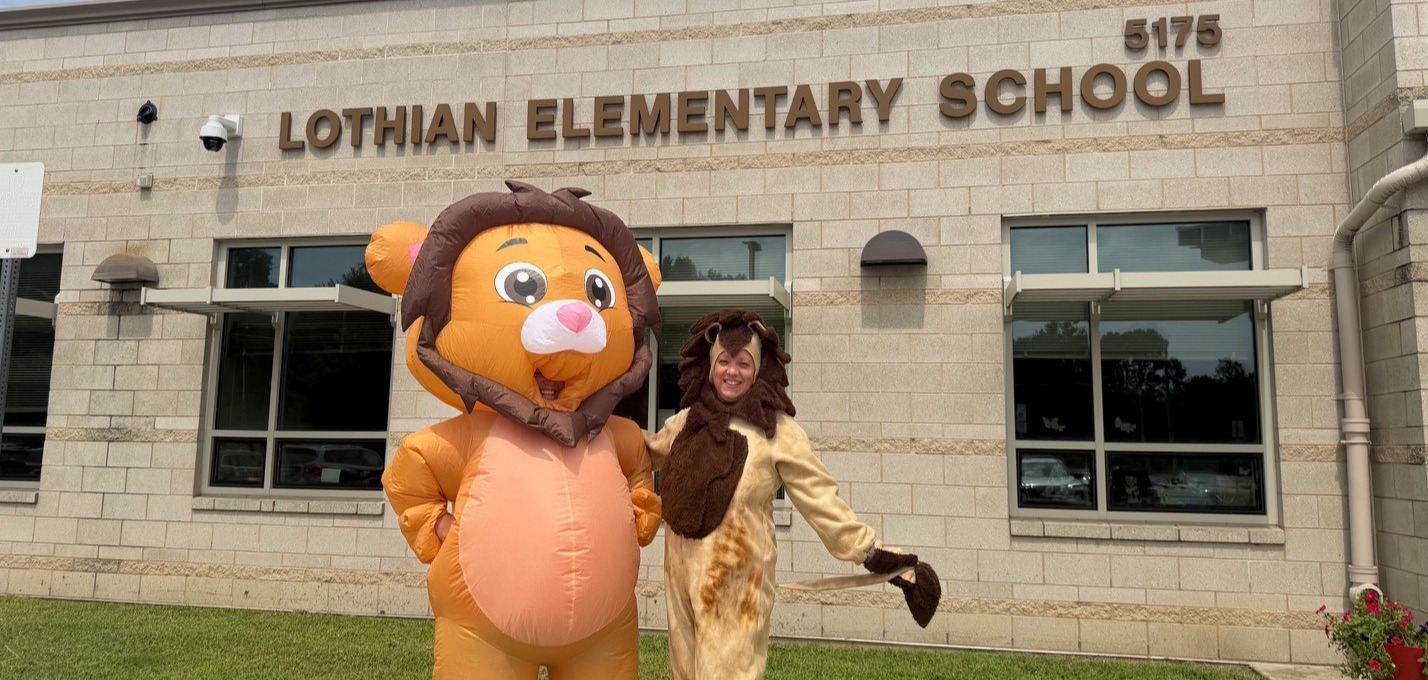 Two individuals dressed in lion costumes stand in front of Lothian Elementary School. One wears a larger lion costume, and the other a smaller one.