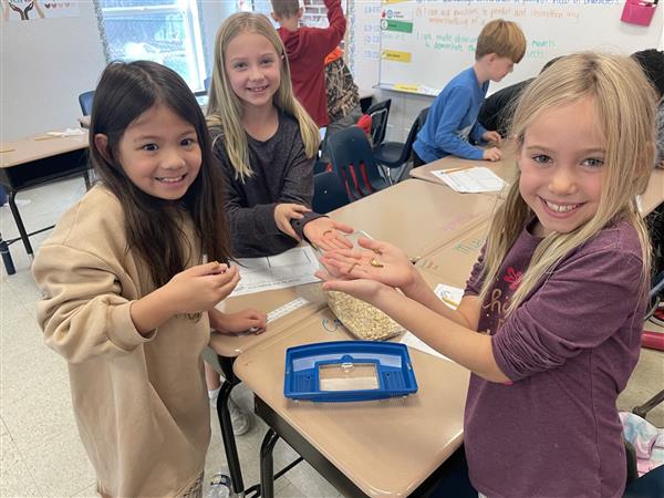 three students are standing around a pod of desks and holding worms