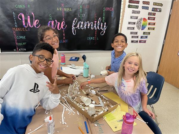 four students work together on a project involving cardboard, paper cups and straws