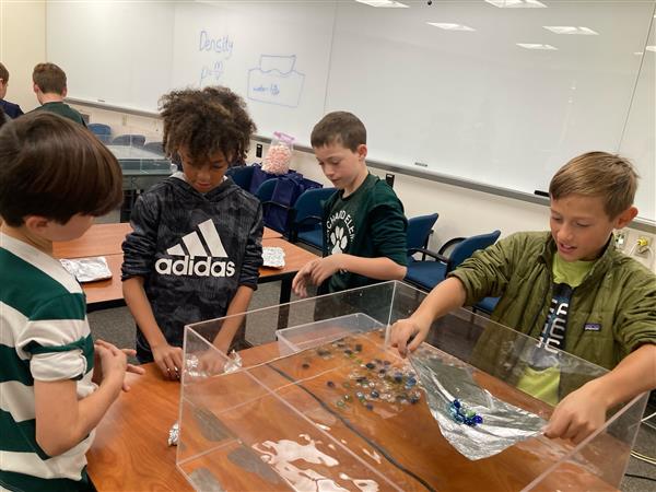 four students are grouped around a large water tank and appear to be working on a science experiment involving submerging marbles in the tank