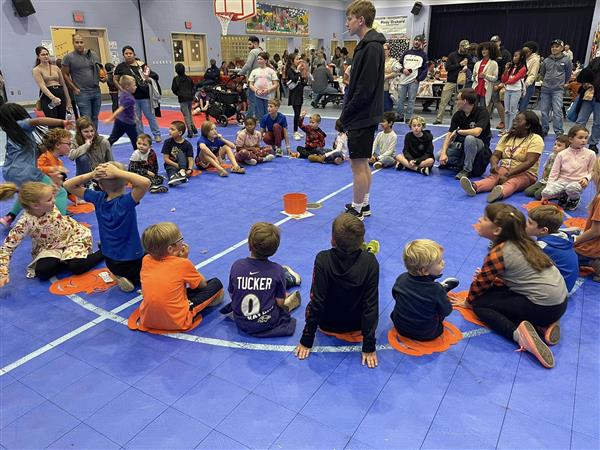 a larger group of students sit in a circle on a blue floor mat 